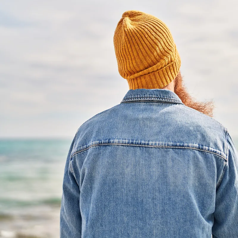 A person in a mustard beanie and denim jacket, seen from behind, looking out to sea