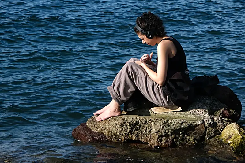 A woman with headphones sitting on a rock by deep blue water, quietly absorbed