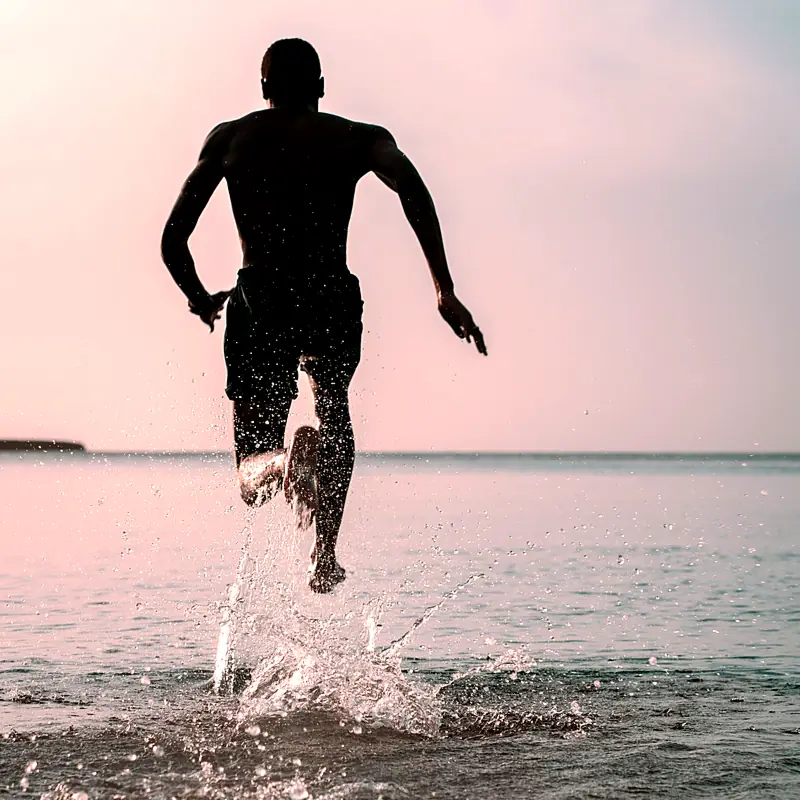 A man running into the sea at dusk, seen from behind, water splashing around his feet